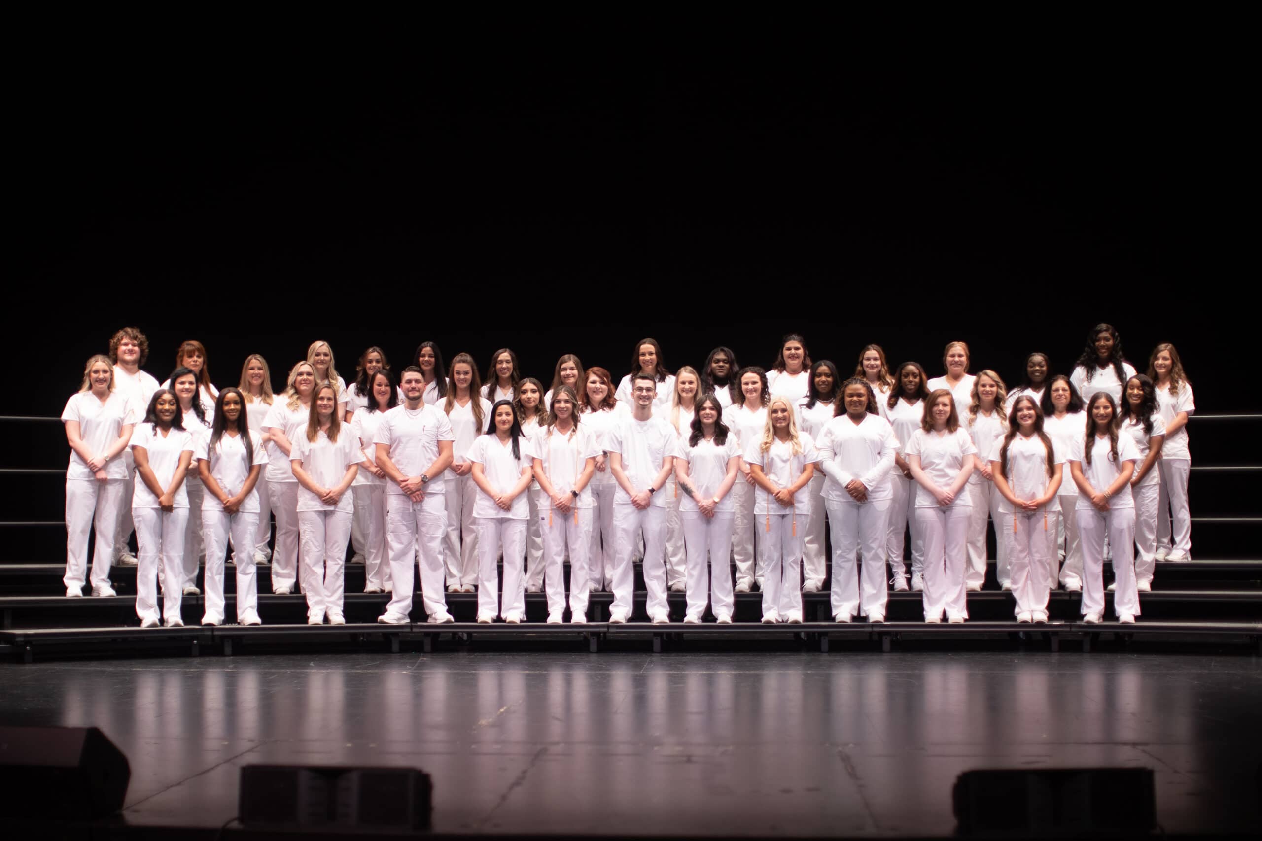 Four rows of men and women in white scrubs on stage for ADN Nursing Program Pinning Sprlng 2022