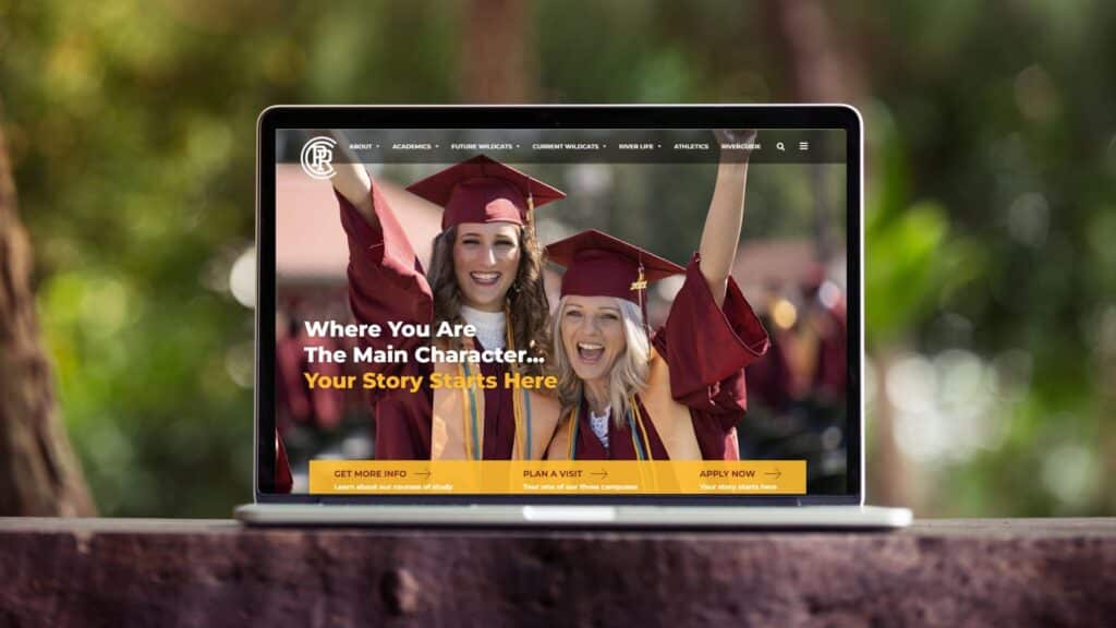Laptop sits on ledge with blurred greenery in the background. Screen shows PRCC website with 2 young women in caps and gowns at graduation smiling with arms raised. Text reads: Where you are the main character... Your story starts here.