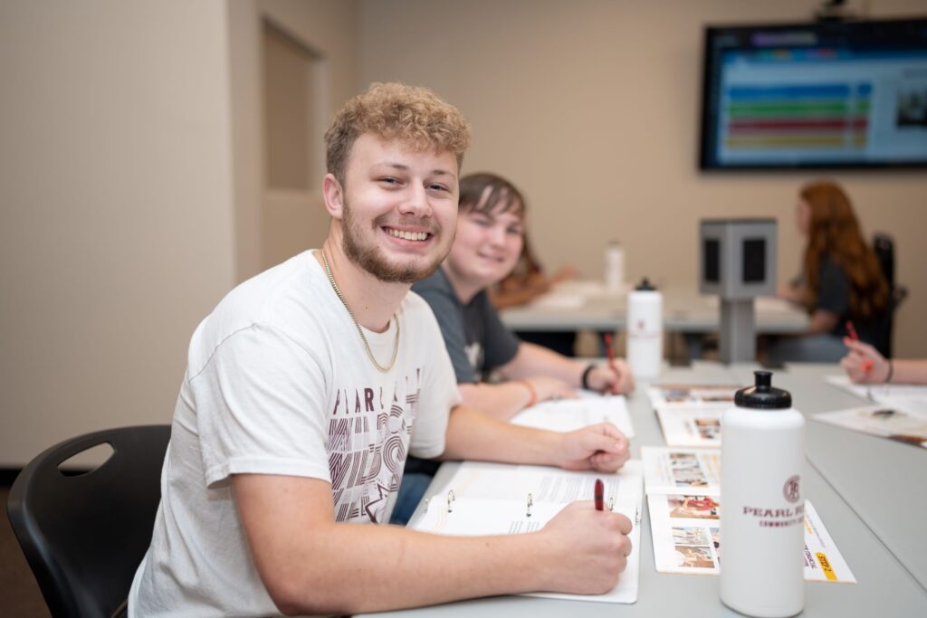 Male college student wearing light colored t-shirt smiles during a class. Other college students in the background.