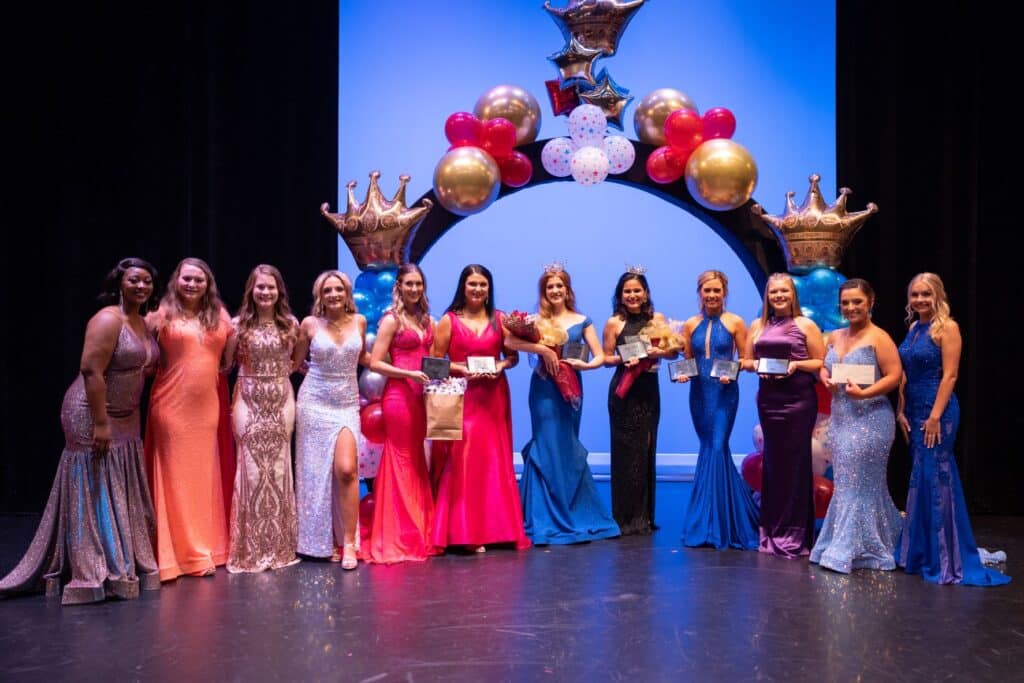 Twelve young women in evening gowns stand in a line with a balloon arch behind them.
