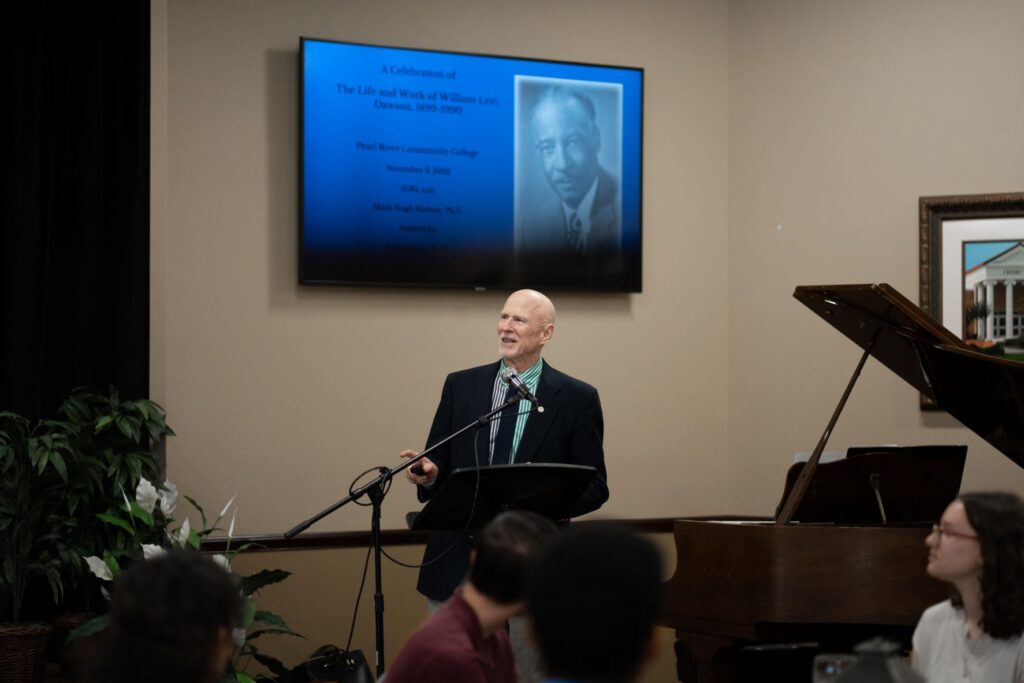 Dr Mark Malone speaks by a microphone while students watch. Baby grand piano and screen with image of William Levy Dawson on it is behind him.
