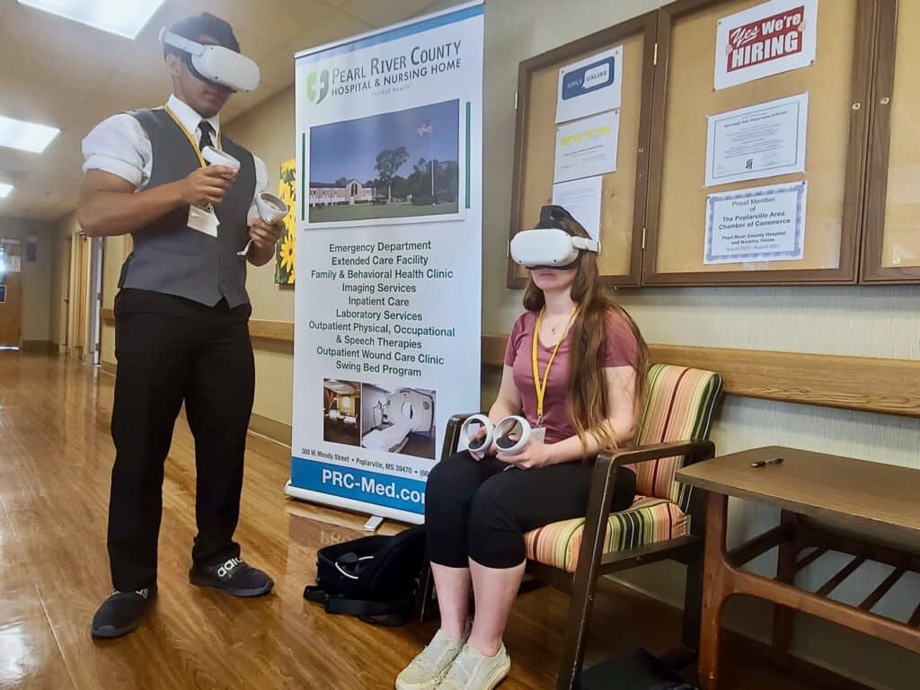 Young man stands while young woman sits. Both are wearing a VR headset. Large banner behind them for Pearl River County Hospital and Nursing Home.