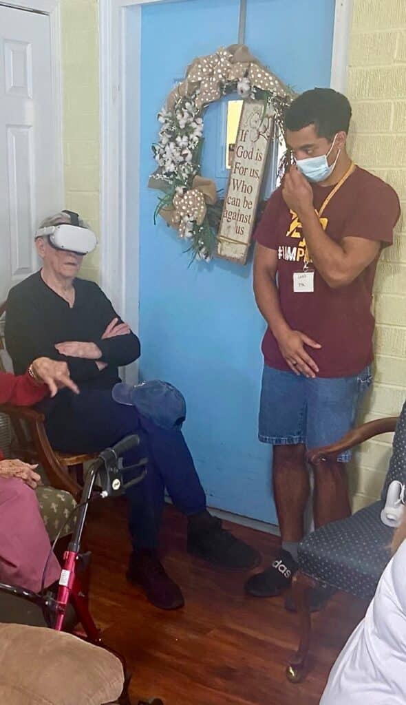 Young man wearing mask observes nursing home resident using VR headset. Large wreath in background.