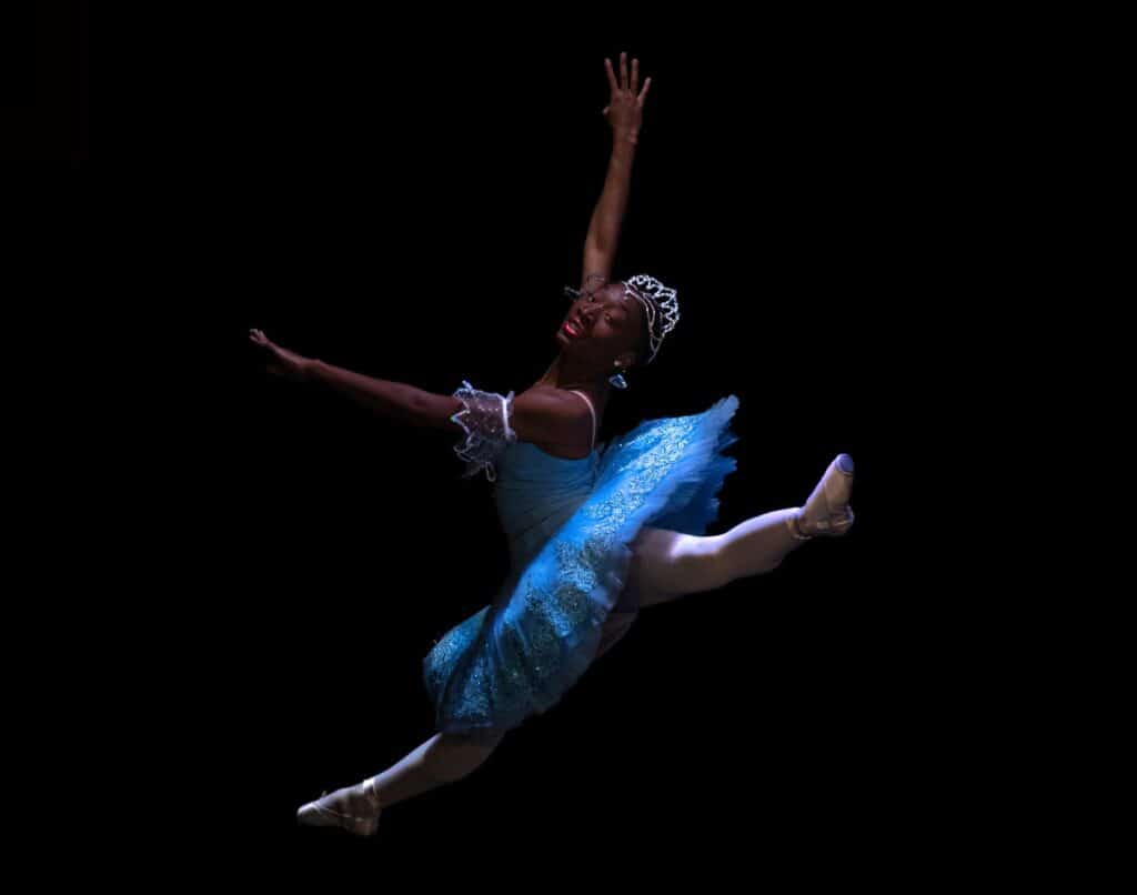 Teenaged girl dressed in blue tutu and tiara does a jumping split.