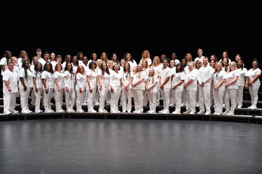 58 men and women in white scrubs stand on stage.