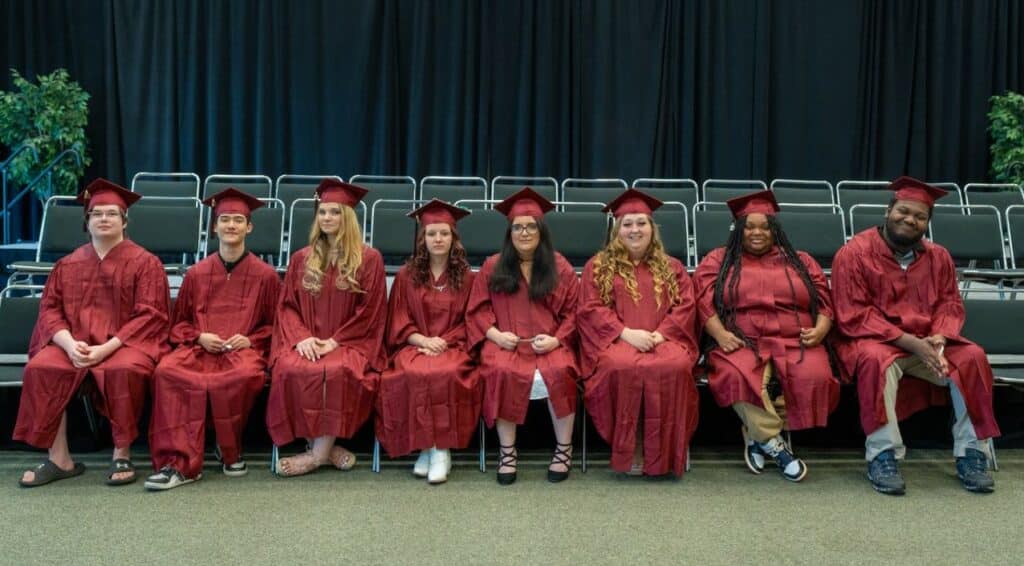 Eight young men and women wearing maroon graduation caps and gowns sit in a row.