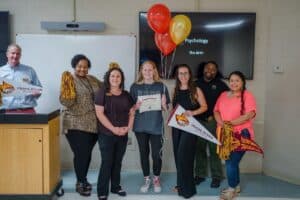 An older man stands near six women of different ages inside a classroom. Balloons and an pendant for Pearl River Community College are held by one lady with a student holding a certificate.