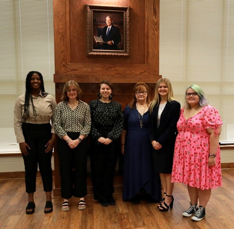 Five students and one faculty member stand in front of fireplace.