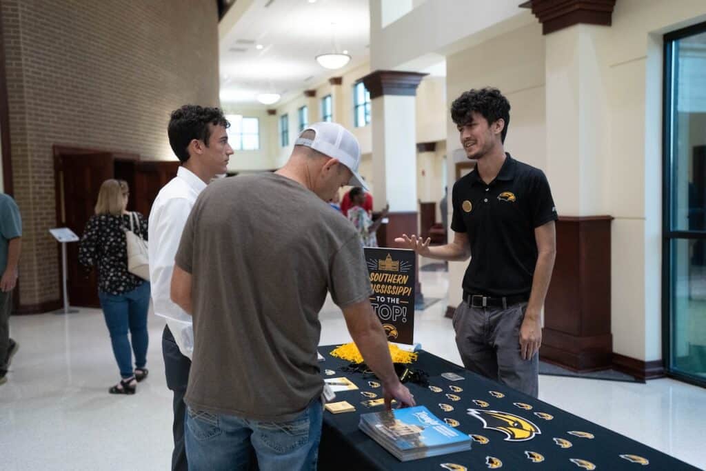Student, parent, and recruiter stand at display table in lobby space.