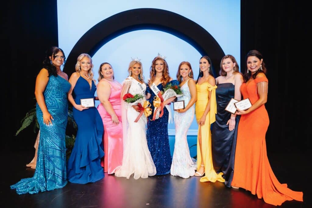 Nine women in evening gowns stand on stage.