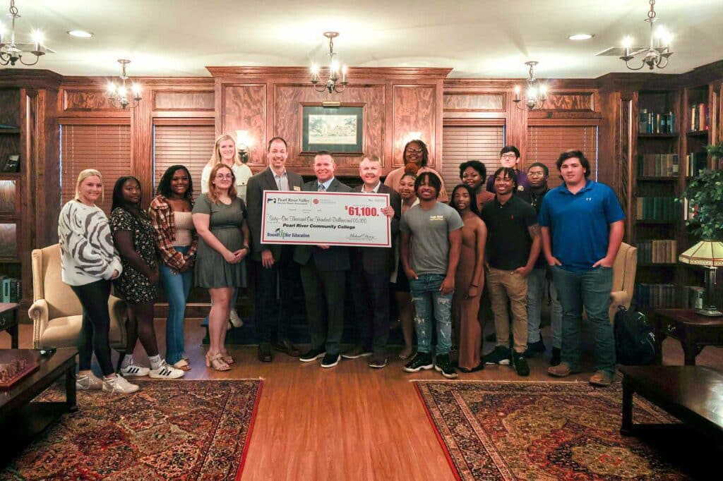 Group of 14 students with 3 adults stand in front of fireplace in wood-paneled library with large check being held.