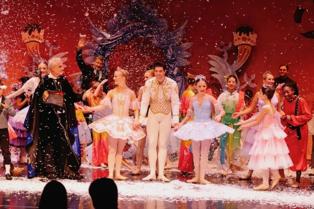 Cast members of The Nutcracker stand on stage with snow coming down.