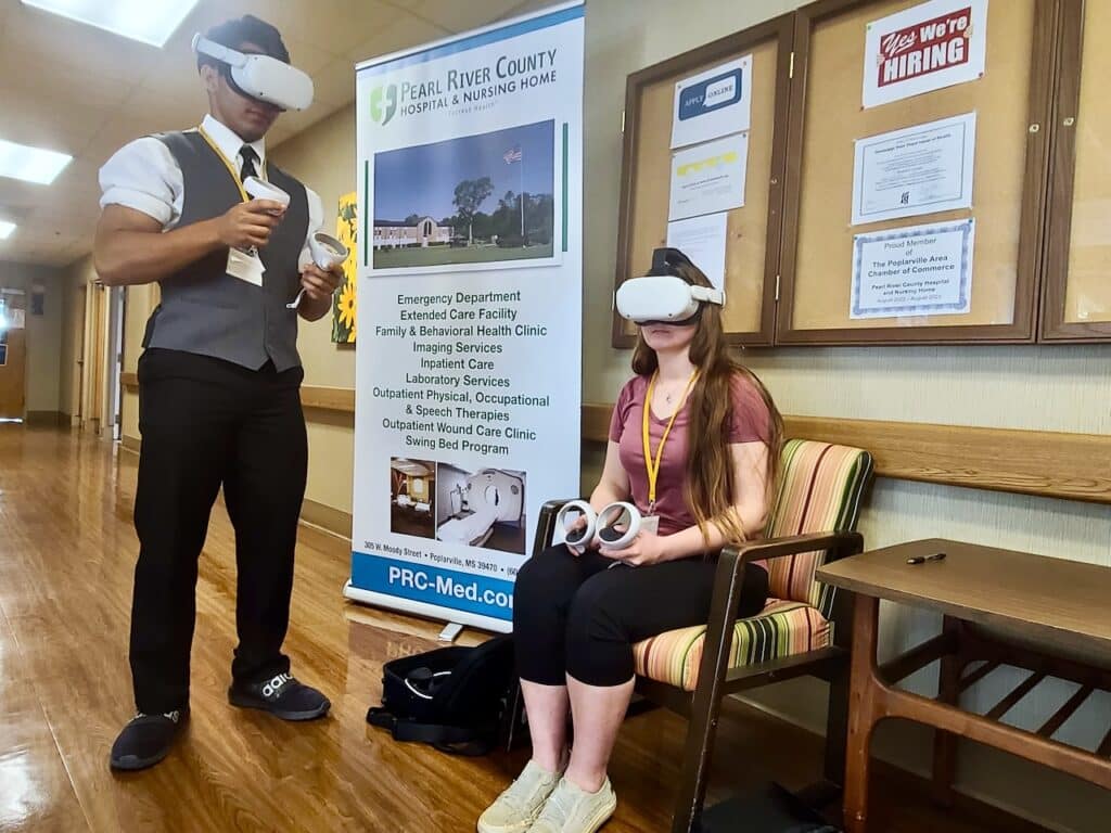 Young man in vest and black pants stands while young woman sits. Both are wearing VR goggles