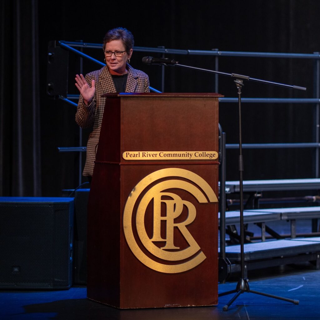 Woman wearing suit speaks while standing behind a podium with PRCC emblem.