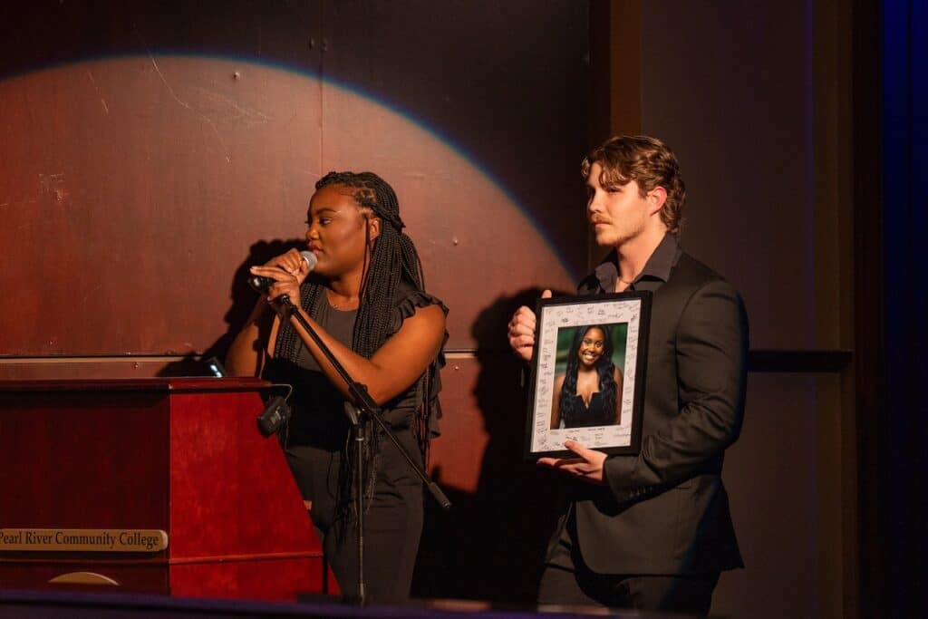 Young woman speaks into microphone while young man holds framed photo.