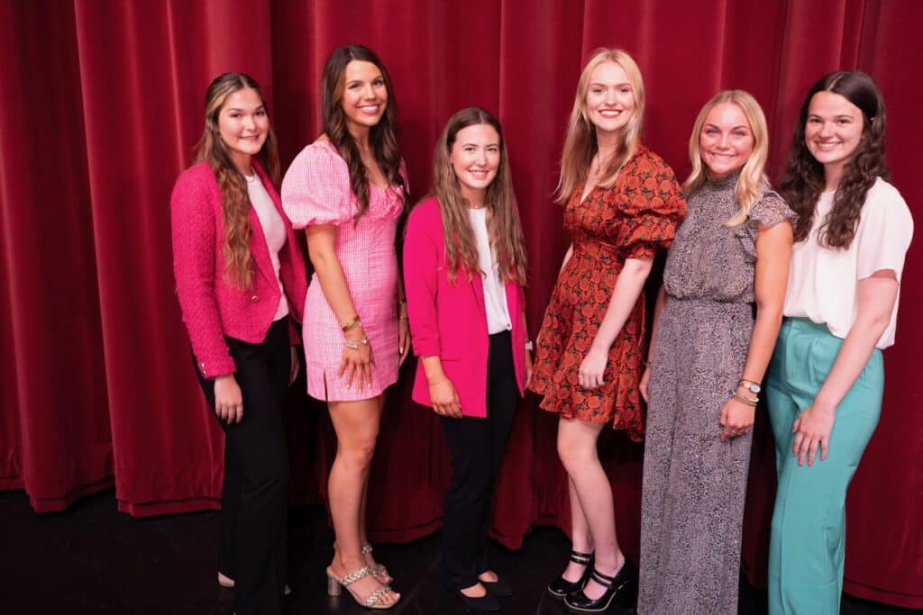 Six smiling young women stand in line on stage.