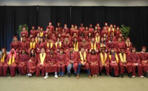 65 men and women wearing maroon caps and gowns sit and stand in rows.