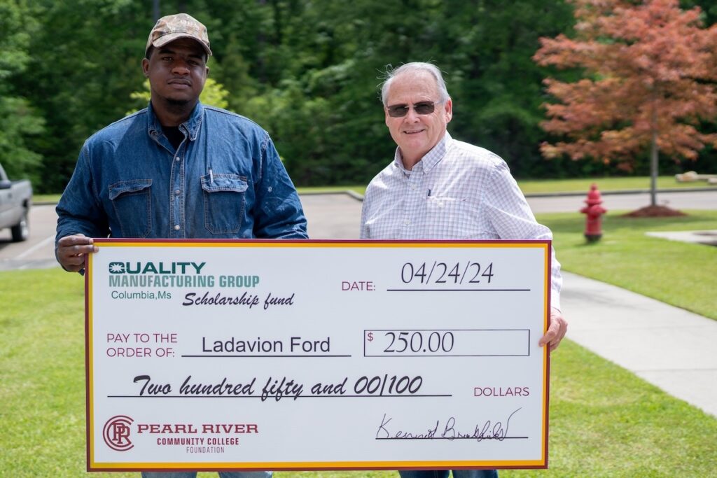 Young man with ball cap holds oversized check with older man wearing sunglasses. Trees and grass in the background.