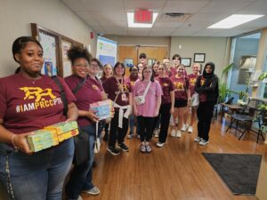Sixteen young adults wearing maroon shirts smile with some holding puzzles and games.