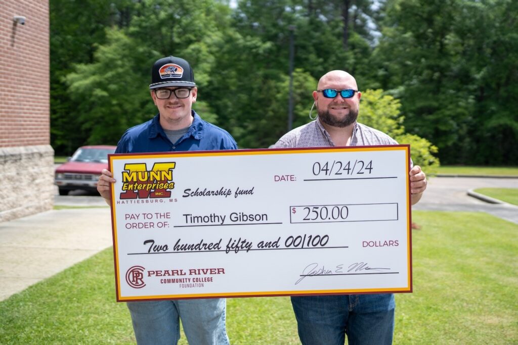 Young man wearing ball cap and another man hold oversized check with trees in the background.