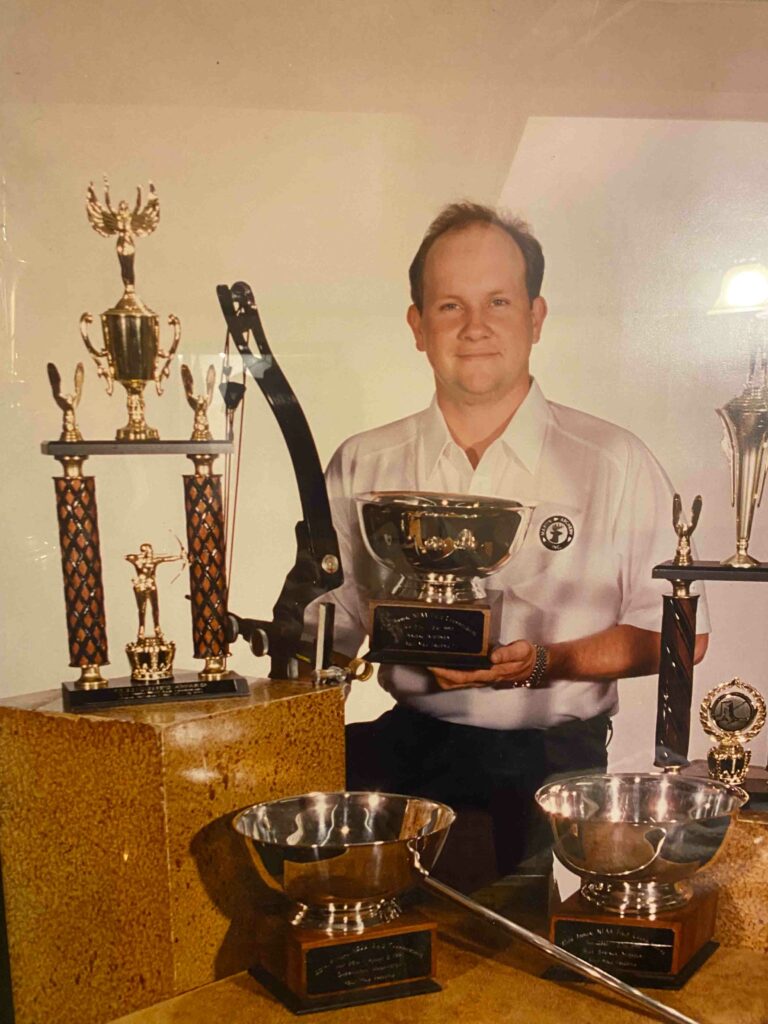 Man holds award plaque with trophies around him.